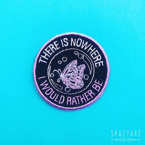 May include: A round black embroidered patch with purple stitching. The patch features a butterfly and the text "There is nowhere I would rather be."