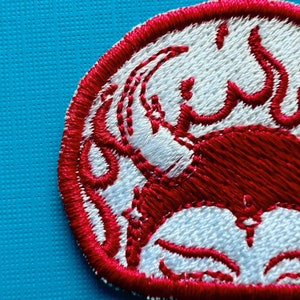 Pu&ograve; includere: Toppa ricamata circolare con un disegno rosso su sfondo bianco, bordata di rosso. Il disegno sembra essere un granchio stilizzato. La toppa ha un aspetto strutturato.