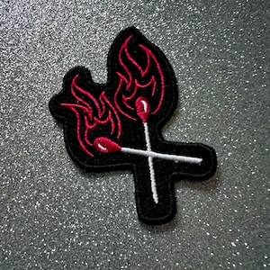 May include: Black and red embroidered patch of two crossed matchsticks with flames. The matchsticks are white and the flames are red.