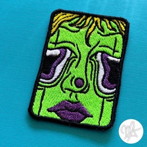 May include: Embroidered patch featuring a green face with purple lips and eyes, and yellow hair. The patch has a black border and is rectangular in shape. The design is a stylized, cartoonish illustration.