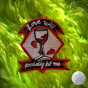 Può includere: Toppa ricamata con un design a calice e la scritta "love will probably kill me". La toppa presenta una combinazione di colori rosso e nero con una grafica a cuore spezzato. La toppa è su uno sfondo verde sfocato.