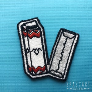May include: Embroidered patch featuring a white rectangular design with red and black accents, and a black border. The patch is open, revealing a detailed interior. The design includes a stylized heart shape. The background is a light blue.