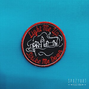 May include: A black and white embroidered patch with red stitching. The patch features a cityscape with the text "Light Me Up Strike Me Down".