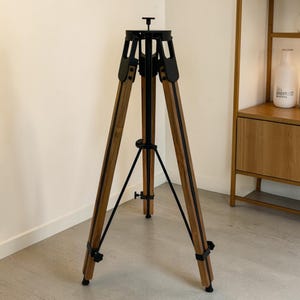 May include: A tripod stand featuring three wooden legs with a natural finish, connected by black metal hardware and a central support brace. The top assembly includes a black metal mounting plate with a small central adjustment knob.