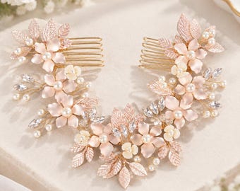 May include: A rose gold hair comb set featuring matte pink floral accents, small white pearls, and clear faceted crystals. The design includes metallic leaves and delicate wire branches, creating a symmetrical arrangement of flowers and foliage.