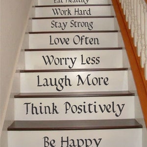 May include: A staircase with white risers featuring black text decals that read: Enjoy Life, Exercise Daily, Eat Healthy, Work Hard, Stay Strong, Love Often, Worry Less, Laugh More, Think Positively, and Be Happy, with dark brown wooden treads.