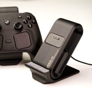 May include: A black charging dock labeled NODE-01 FOR STEAM CONTROLLER with a gray fabric base, positioned next to a black wireless game controller featuring dual analog sticks, a directional pad, and a central Steam logo button.
