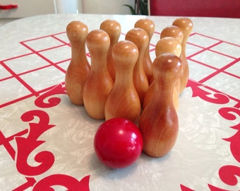 May include: A set of wooden bowling pins arranged in a triangle shape on a white surface with red grid lines. A red bowling ball sits in front of the pins.