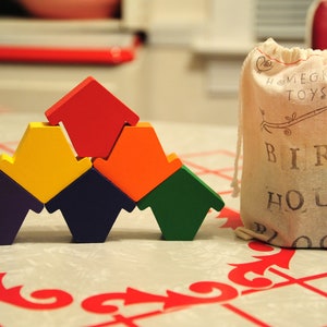 Puede incluir: Un conjunto de bloques de madera de colores con forma de casa para pájaros. Los bloques están dispuestos en forma de pirámide y están etiquetados como "Bird House Blocks".
