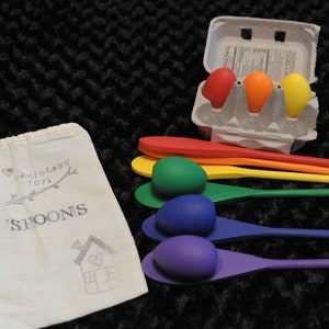 May include: A set of wooden spoons with rainbow colored handles and matching colored eggs. The spoons are arranged in a rainbow pattern with the purple spoon on the bottom and the red spoon on top. The eggs are in a cardboard egg carton with the words "Homegrown Toys" on the side. The spoons and eggs are on a black fuzzy surface.