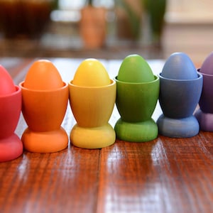 May include: Six wooden egg cups in a rainbow of colors, from red to purple, with a wooden egg in each cup.