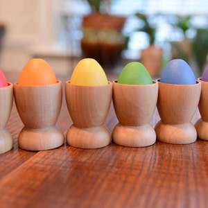 Może przedstawiać: Sześć drewnianych stojaków na jajka z kolorowymi jajkami w środku. Jajka są czerwone, pomarańczowe, żółte, zielone, niebieskie i fioletowe.