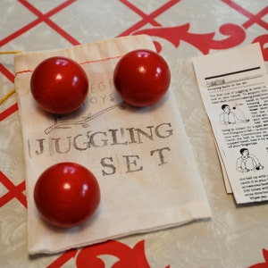 Może przedstawiać: Biała torba płócienna z nadrukiem "Juggling Set". Wewnątrz torby znajdują się trzy czerwone piłki do żonglowania. Obok torby leży kartka papieru z instrukcjami, jak żonglować.