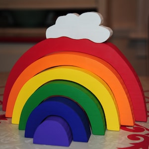 May include: A rainbow-colored wooden arch toy with a white cloud on top. The arch is made of seven different colored pieces, each with a slightly different shape. The toy is perfect for toddlers and young children to play with.