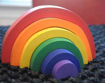 May include: A rainbow-colored wooden arch toy with seven different colored pieces, including red, orange, yellow, green, blue, indigo, and violet.