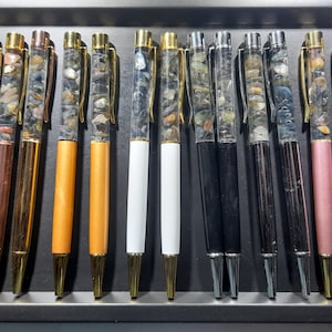 Könnte beinhalten: Eine Schale mit 14 Stiften mit durchsichtigen Fässern, die mit bunten Steinen gefüllt sind. Die Stifte haben goldene Akzente und verschiedene farbige Fässer, darunter Kupfer, Braun, Weiß, Schwarz und Rosa.