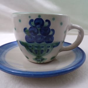 Puede incluir: Un juego de taza y platillo de cerámica blanca con un diseño floral azul y verde. La taza tiene una forma redondeada y un asa. El platillo tiene un borde azul.
