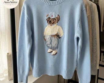 May include: A light blue long-sleeve crewneck sweater featuring a central graphic of a brown teddy bear wearing a white hat, a yellow sweater, and light blue pants. The sweater has a ribbed collar, cuffs, and hem, with a Polo label at the neck.