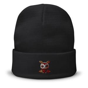 Op de afbeelding: Een effen zwarte muts met omslag en een kleine, centraal geplaatste borduursel van een gestileerde mot met rode, oranje en witte draaddetails. De mot heeft grote, donkere ogen en kleine vleugels op de voorzijde van de omslag.