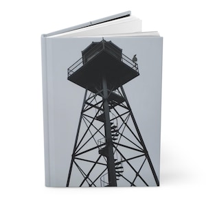 May include: A hardcover journal featuring a black and white photograph of a tall, metal watchtower with a spiral staircase and a small observation cabin at the top, set against a plain, light gray sky background.