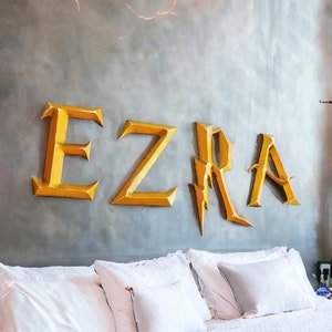 May include: A bedroom with a white bed and a gray wall with the name "EZRA" written in gold letters. The letters have a lightning bolt design.