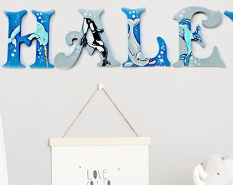 May include: A nursery decor set featuring the name "HALEY" in blue and gray wooden letters with a sea life theme. Each letter is decorated with a different sea creature, including a dolphin, whale, and narwhal. The letters are displayed on a white wall.