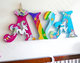 May include: A set of three wooden letters spelling "AVA" painted in bright colours with a unicorn theme. Each letter features a rainbow, stars, and a unicorn. The letter "A" at the beginning of the name has a unicorn horn on top.