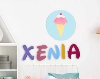 Könnte beinhalten: An einer weißen Wand sind bunte Holzbuchstaben mit dem Namen "XENIA" angebracht. Darüber befindet sich ein hellblauer Kreis mit einer Eiswaffel-Illustration. Ein Regal und ein Bettrahmen sind ebenfalls sichtbar.