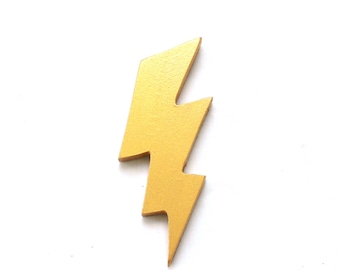 May include: A gold-colored lightning bolt cutout made of wood. The lightning bolt has a smooth, matte finish and a sharp, angular shape. The logo for The Fairy Paintbox, featuring text and a butterfly icon, is visible in the bottom left corner.
