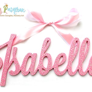 May include: Pink wooden name sign with the name "Isabella" spelled out in cursive letters. The letters are covered in small, clear rhinestones. A pink satin ribbon with a bow is attached to the top of the sign.