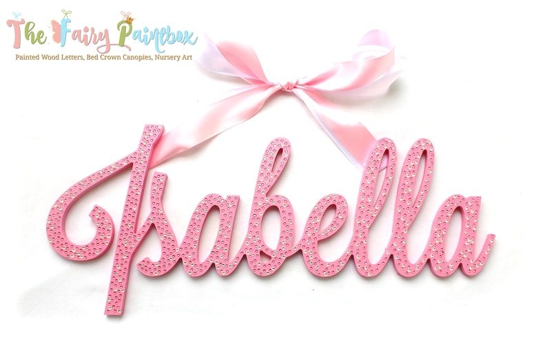 May include: Pink wooden name sign with the name "Isabella" spelled out in cursive letters. The letters are covered in small, clear rhinestones. A pink satin ribbon with a bow is attached to the top of the sign.