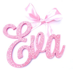 May include: Pink wooden name sign with the name "Ella" spelled out in cursive. The sign is decorated with small, clear rhinestones and has a pink satin ribbon attached to the top.