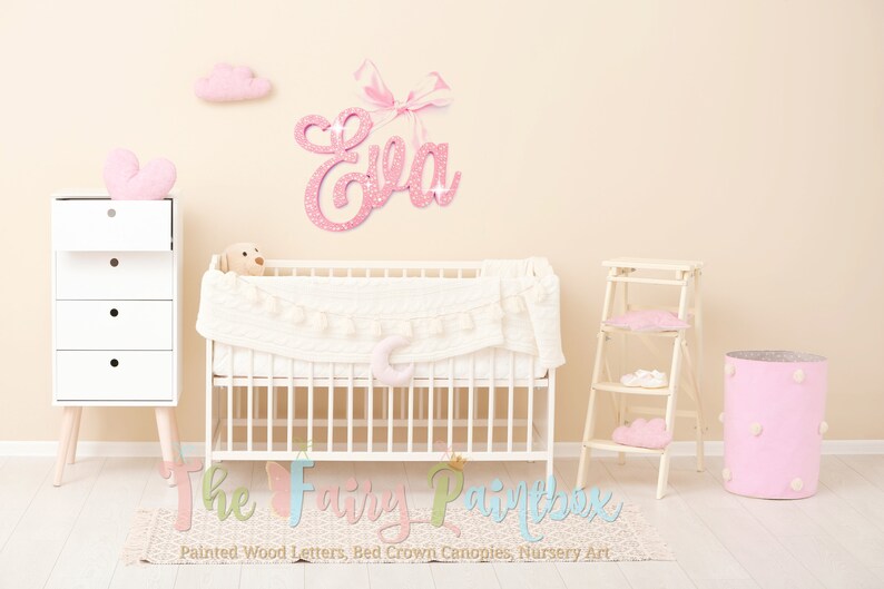 May include: Pink glitter name sign with a bow, spelling the name "Eva", hanging on a light beige wall above a white crib with a white and pink blanket. The crib is in a nursery with a white dresser, a pink storage bin, and a white step stool.