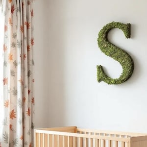 May include: A large, three-dimensional letter S covered in vibrant green preserved moss. The letter is mounted on a plain white wall above a light-colored wooden crib, positioned next to a curtain featuring a pattern of orange and gray leaves.