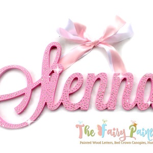 May include: Pink wooden name sign with the name "Elena" spelled out in cursive letters. The letters are covered in rhinestones and have a sparkly finish. A pink and white ribbon is tied at the top of the sign.