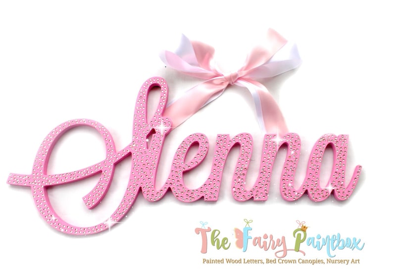 May include: Pink wooden name sign with the name "Elena" spelled out in cursive letters. The letters are covered in rhinestones and have a sparkly finish. A pink and white ribbon is tied at the top of the sign.