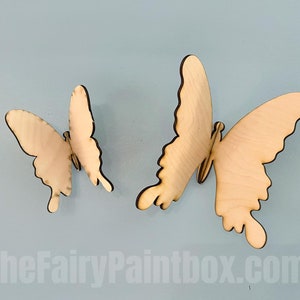 May include: Two wooden butterflies, one larger than the other, are attached to a light blue wall. The butterflies are unpainted and have a natural wood color.