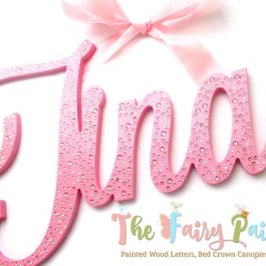 May include: Pink wooden letters spelling the name "Jina" with a pink bow on top. The letters are decorated with small, clear rhinestones. The letters are on a white background. The text "The Fairy Paintbox" is at the bottom of the image.