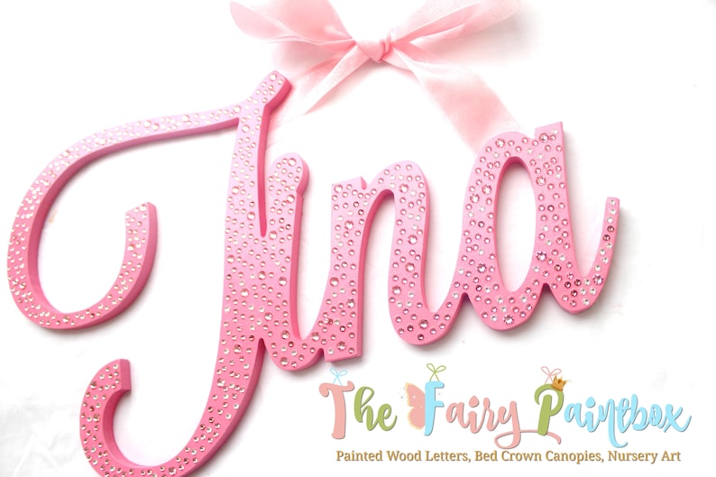 May include: Pink wooden letters spelling the name "Jina" with a pink bow on top. The letters are decorated with small, clear rhinestones. The letters are on a white background. The text "The Fairy Paintbox" is at the bottom of the image.
