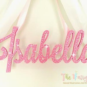 May include: Pink glitter wooden name sign with the name "Isabella" in cursive script. The sign is hanging from a white ribbon.