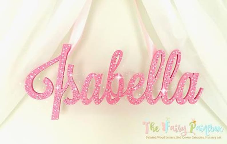 May include: Pink glitter wooden name sign with the name "Isabella" in cursive script. The sign is hanging from a white ribbon.