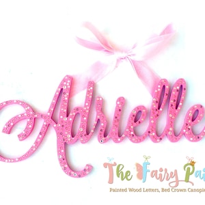 May include: Pink wooden letters spelling the name "Adrielle" with rhinestones. The letters are attached to a pink ribbon.