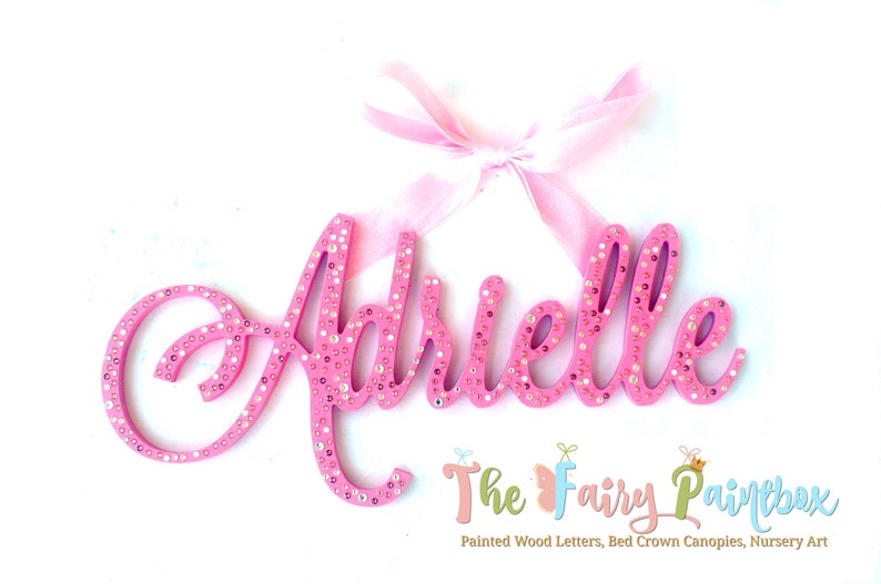 May include: Pink wooden letters spelling the name "Adrielle" with rhinestones. The letters are attached to a pink ribbon.