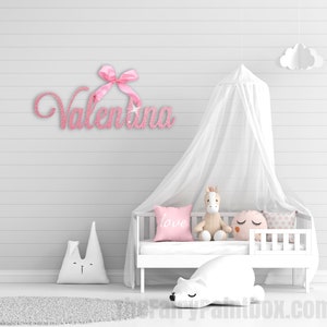 May include: Pink glitter name sign with a pink bow, spelling out the name "Valentina". The sign is hanging on a white wall above a white crib with a white canopy and a stuffed animal.