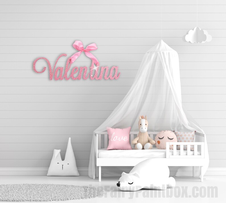 May include: Pink glitter name sign with a pink bow, spelling out the name "Valentina". The sign is hanging on a white wall above a white crib with a white canopy and a stuffed animal.