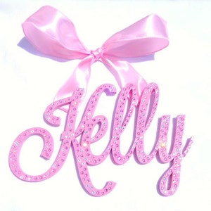 May include: Pink wooden name sign with the name "Kelly" spelled out in cursive script. The sign is decorated with pink rhinestones and has a pink satin bow attached to the top.