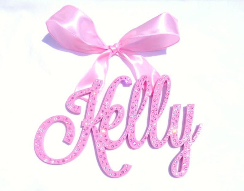 May include: Pink wooden name sign with the name "Kelly" spelled out in cursive script. The sign is decorated with pink rhinestones and has a pink satin bow attached to the top.