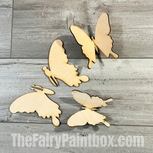 May include: Four unfinished wooden butterfly shapes. The butterflies are in various sizes and positions on a light gray wood background.
