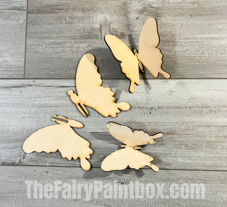 May include: Four unfinished wooden butterfly shapes. The butterflies are in various sizes and positions on a light gray wood background.