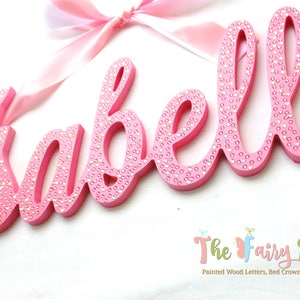 May include: Pink wooden letters spelling the name "Isabella" with small white rhinestones embedded in the paint. The letters are in a cursive font and are hanging from a pink ribbon.